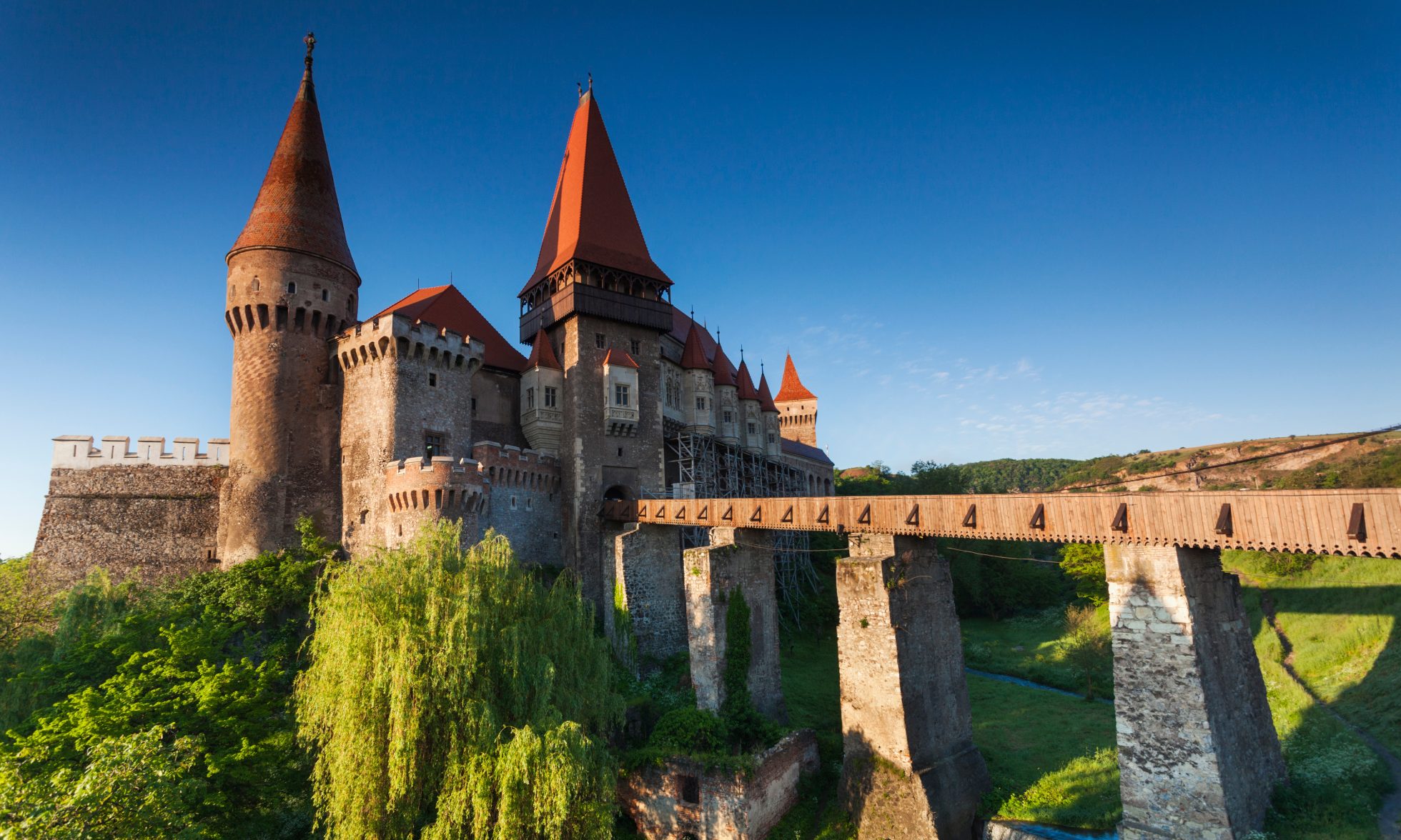The photo shows Corvin Castle in Transylvania, Romania. The castle is surrounded by mountains and trees.