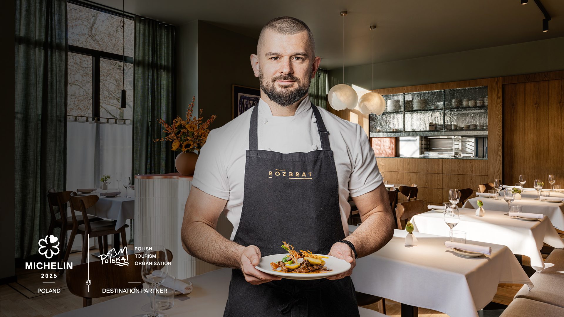 Szef kuchni Rozbrat 20 Bartosz Szymczak trzymający danie. Wnętrze restauracji w tle
