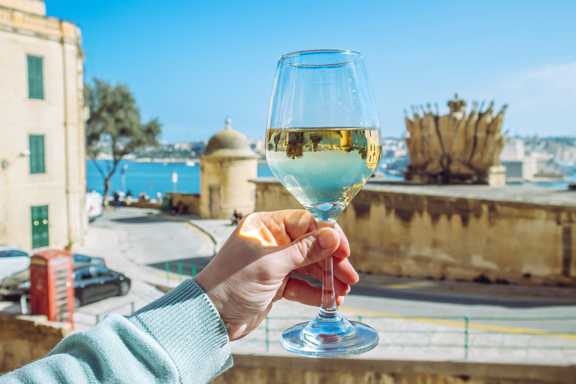 Selektywne skupienie na kobiecie trzymającej kieliszek białego wina w pięknym mieście Valletta na Malcie. Tło z błękitnym niebem i morzem w słoneczny dzień. Koncepcja wycieczki po winnicach.