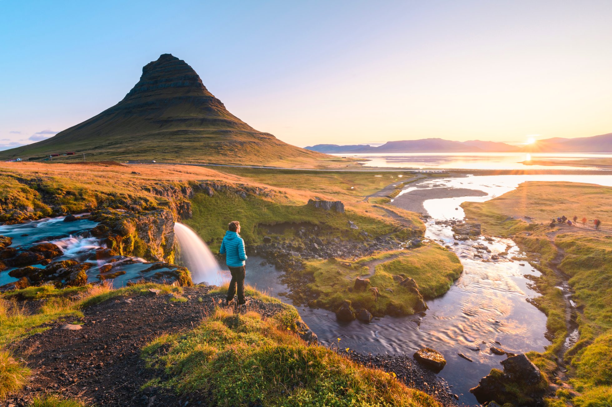 A tourist stands by a waterfall under the Kirkjufell mountain and watches the sunset.