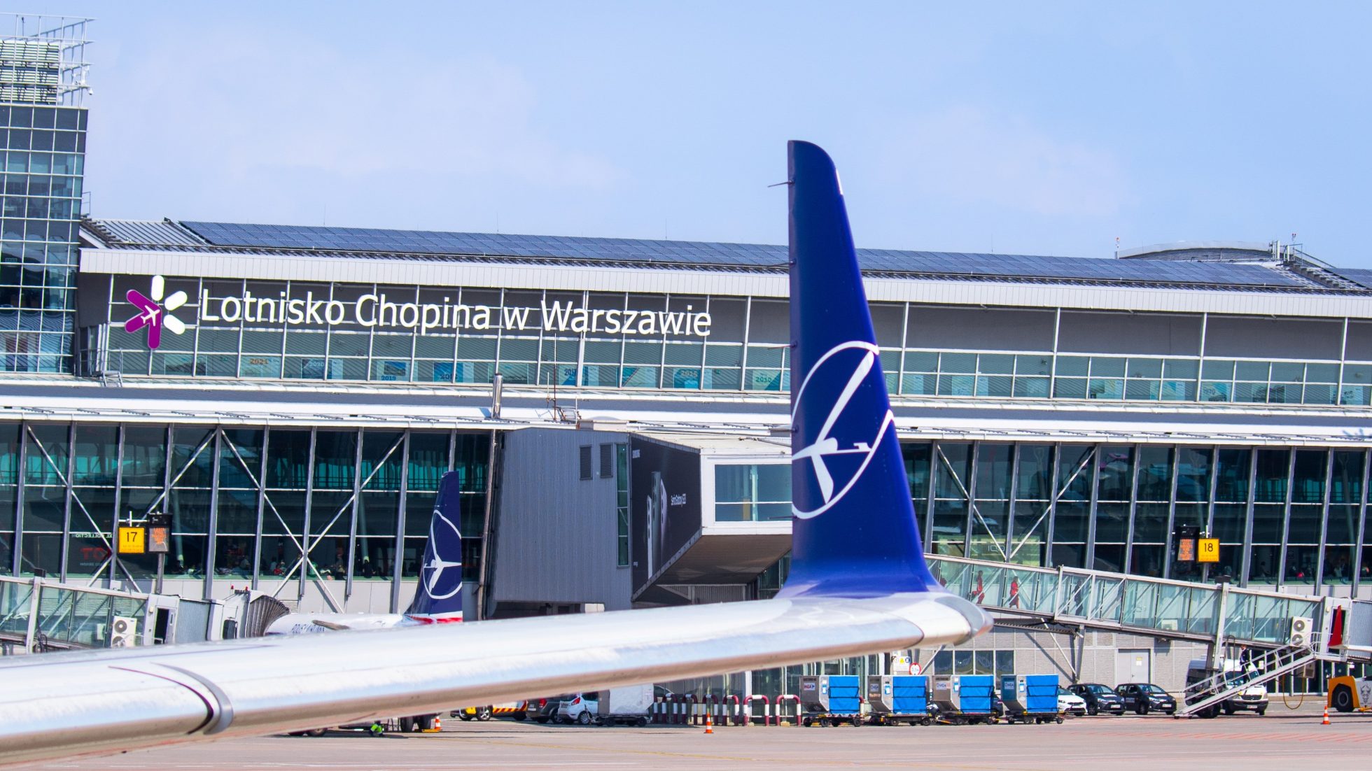 Wing of the Embraer 175 with LOT' Crane logo on the winglet with Chopin Airport terminal in the background on a sunny day