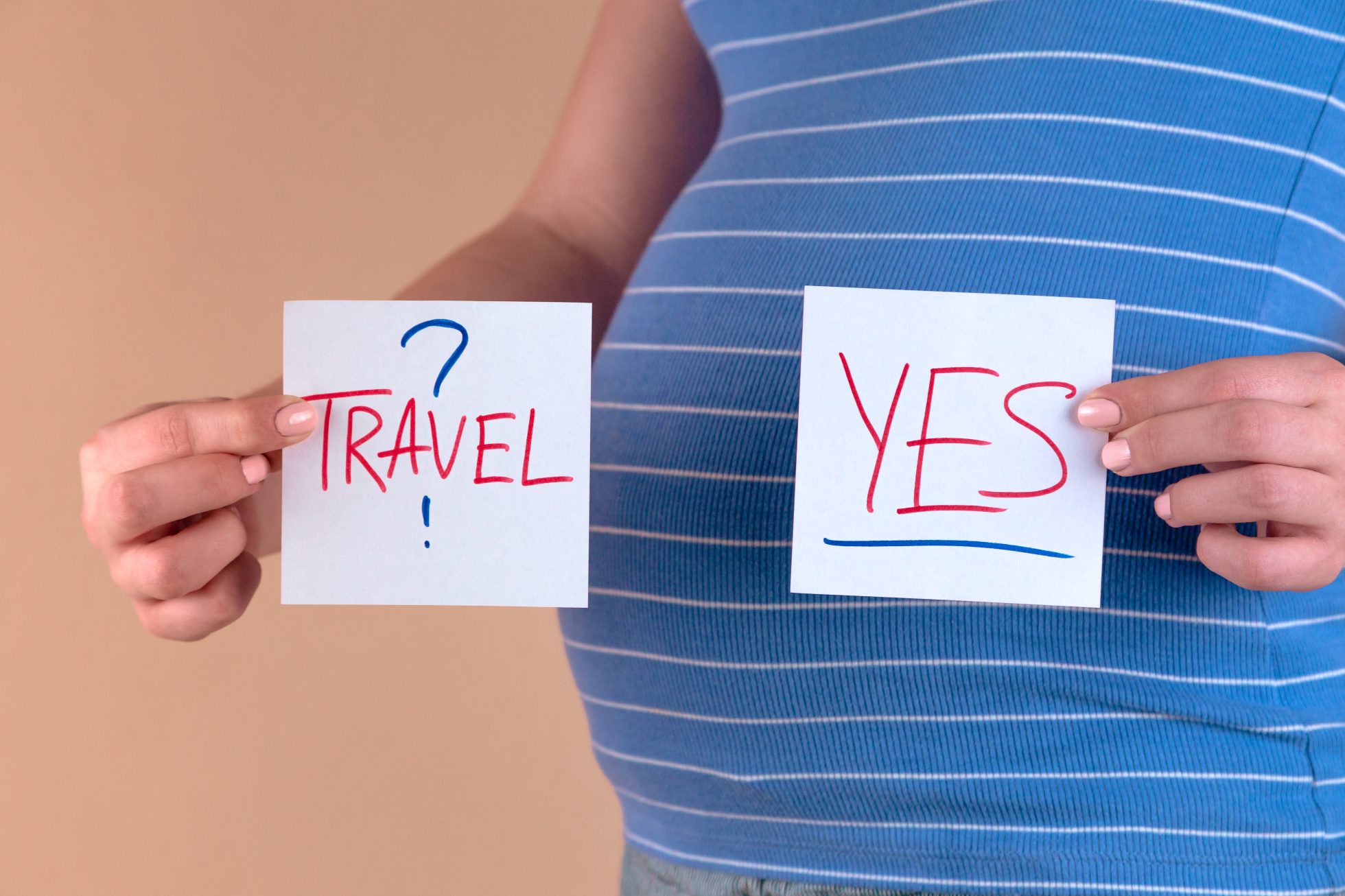 Close-up of the belly of a pregnant woman in a blue T-shirt, who is holding leaflets with the words travel and Yes. The concept of travel during pregnancy, flying on a plane while pregnant