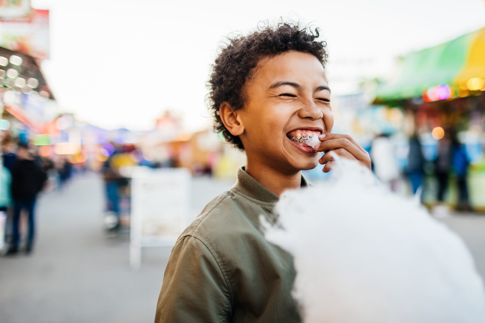 A young boy with braces on his teeth smiling while eating candy floss at the fun fair.