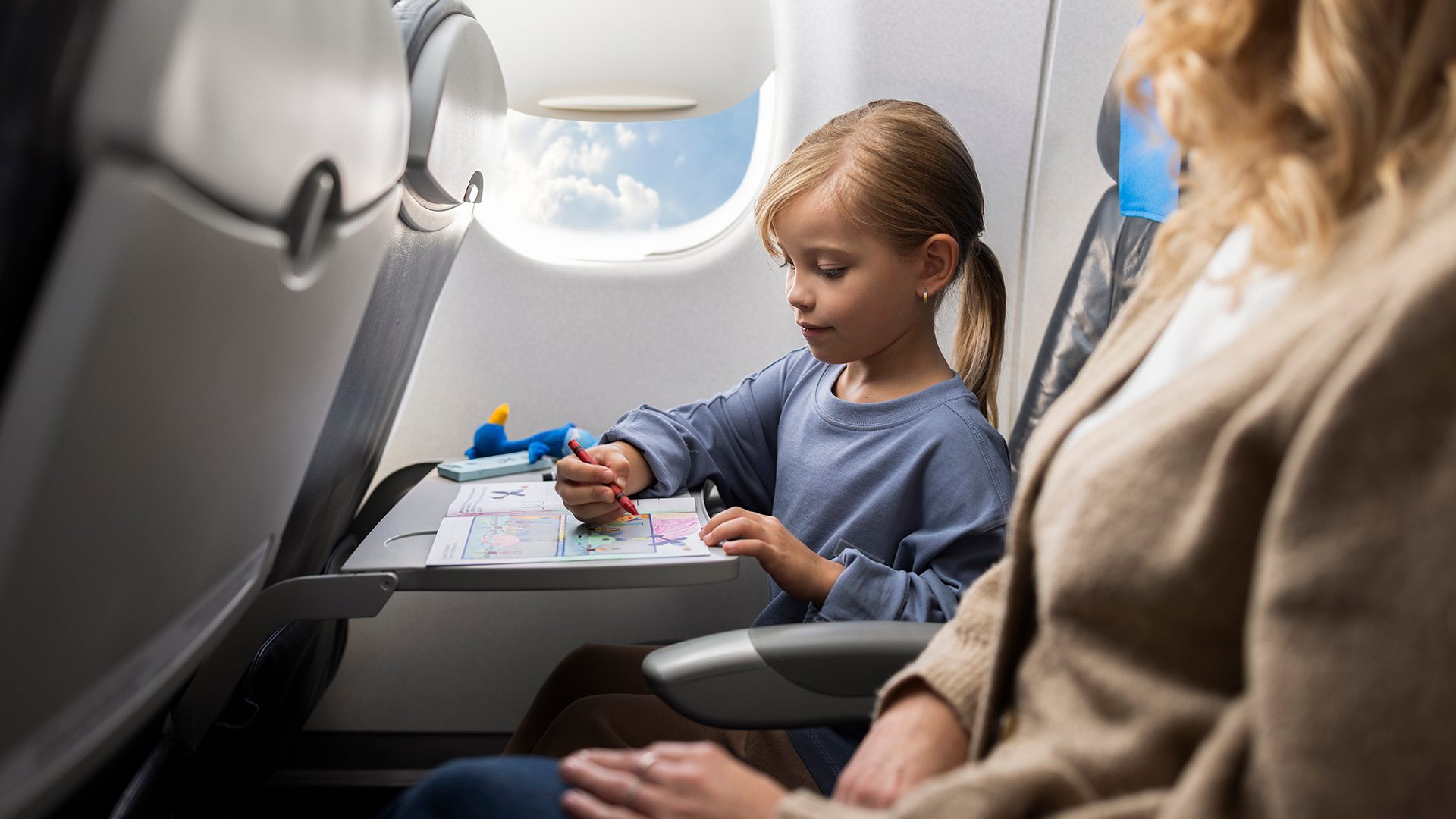 A child onboard an Embraer is coloring in a coloring book, with a Lotek plush toy lying nearby.