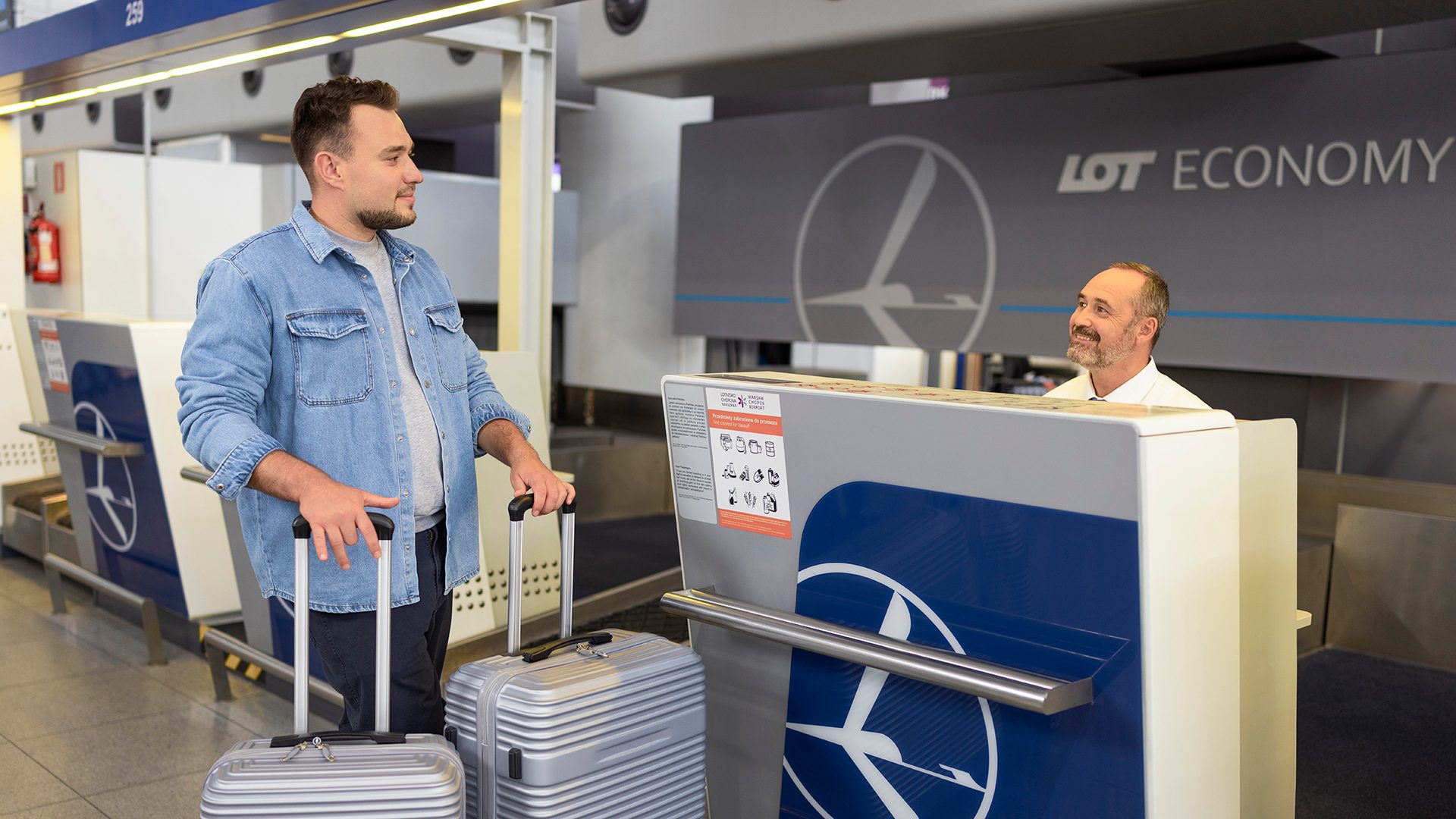 A passenger with two suitcases is greeted by a smiling LOT staff member at the check-in counter.